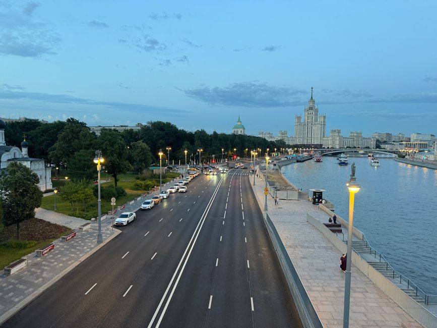 A wide view of the River Thames at dusk, showing an empty multi-lane road with white lane markings in the foreground, lined with evenly spaced streetlights. On the left, there is a park area with gree