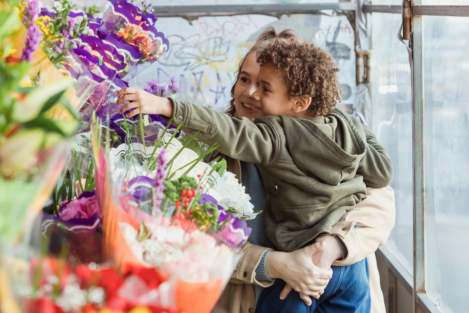 A young girl with curly hair, smiling happily, reaches out to select a floral arrangement in a market stall. She is being supported by an adult, whose arms are wrapped around her waist. The floral dis