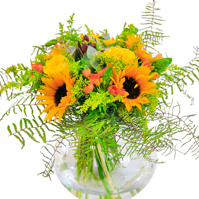 The first impression of this arrangement is a joyous burst of sunlight, with vivid yellow sunflowers and garden roses sharing centre stage, their petals unfolding in layers of golden warmth reminiscent of peak summer in Seven Sisters' Woodberry Down. Sunflowers, with their dark, textured hearts and velvety-yellow rays, exude a sense of optimism and rural charm, while the garden roses spiral open in tighter, more refined blossoms. Scarlet Hypericum berries glitter among abundant foliage, providing playful contrast to the soft, matte petals. Greenery flows outward in wispy sprays-ferns, fillers, and the pale hint of eucalyptus-all tumbling gracefully across the lip of the clear, bowl-shaped glass vase. At the base, the intricate weave of sturdy green stems shimmers through clear water, amplifying the freshness of the entire bouquet. The careful, flowing architecture of each flower and leaf testifies to the expertise of Seven Sisters Florist. This is a bouquet designed to mark joyful milestones: birthdays, anniversaries, or heartfelt congratulations, especially in the thriving, multicultural heart of Seven Sisters-a place where celebration and togetherness are always in season.
